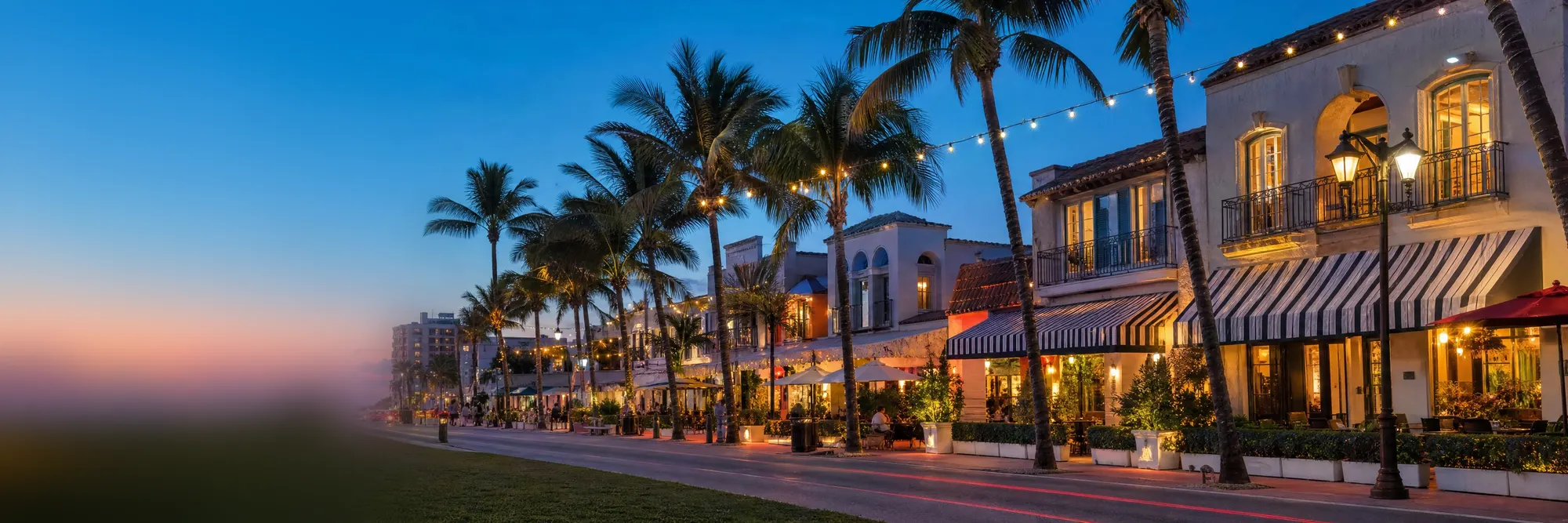 A palm-lined main street at dusk, its cafés lit up.